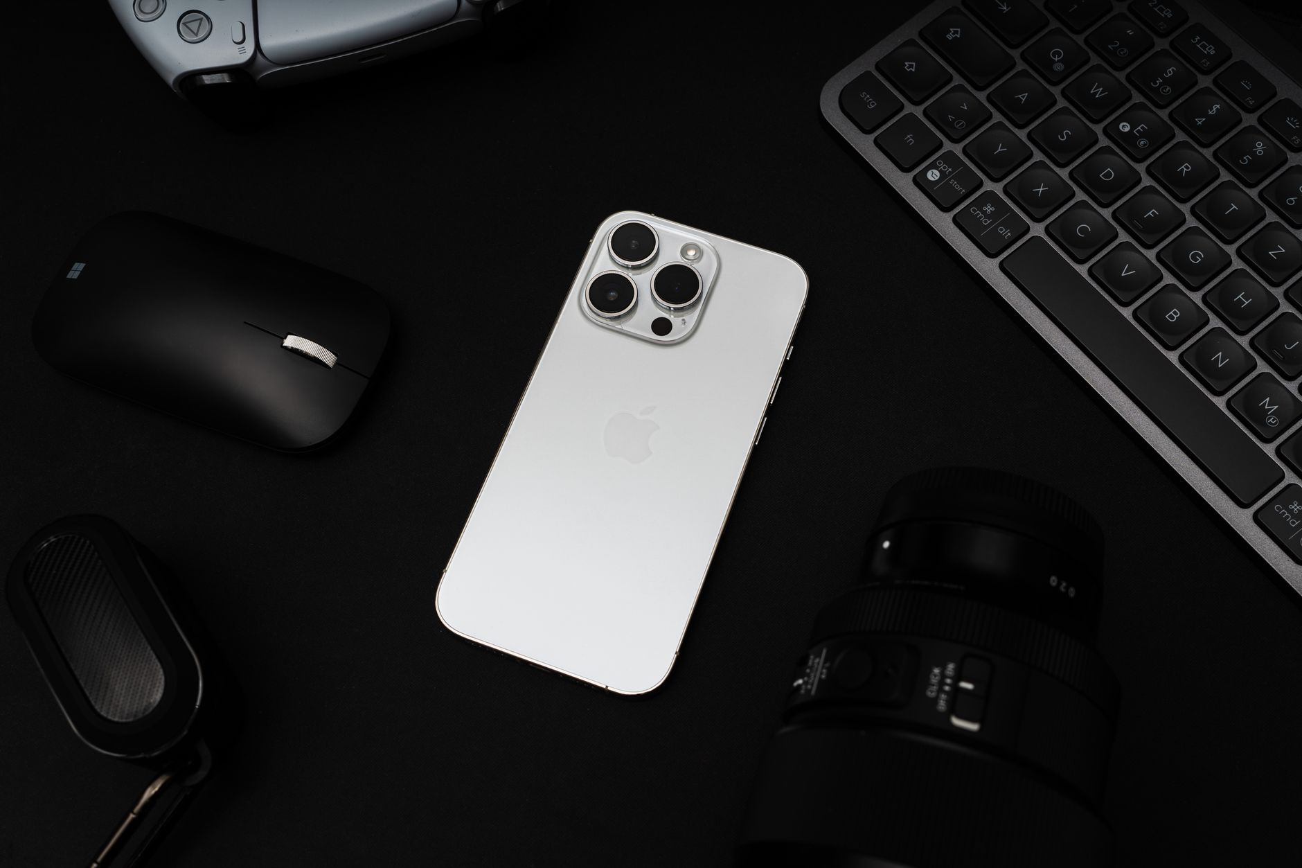 Hvid smartphone med trippel kamera omgivet af gaming-controller, keyboard og trådløse øretelefoner på sort baggrund