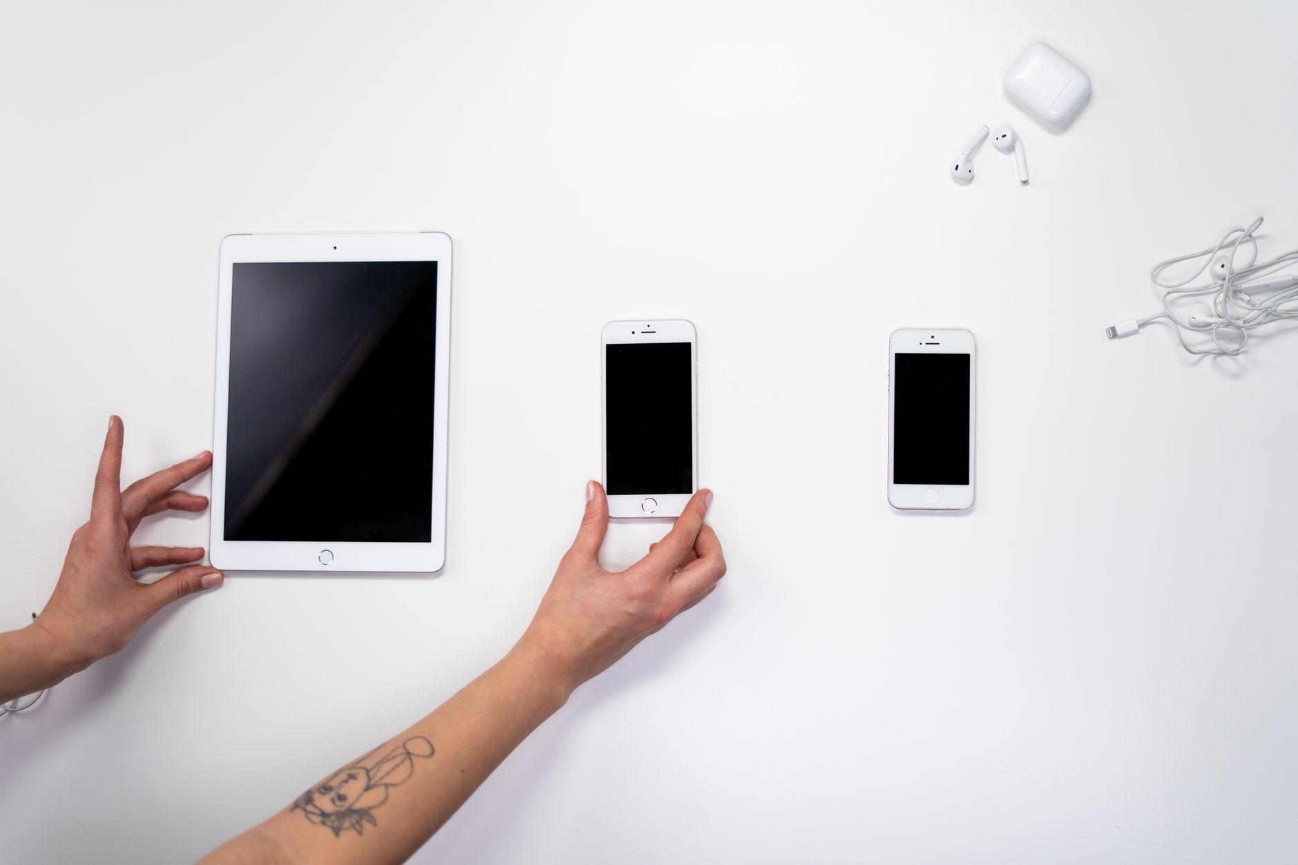 Hænder holder hvid tablet og smartphone ved siden af tilbehør som øretelefoner, opladningskabel og case på hvid baggrund